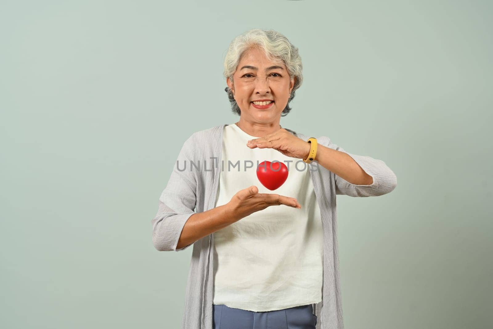 Smiling 60s woman holding red heart over blue background. Health care, medical and charity concept.