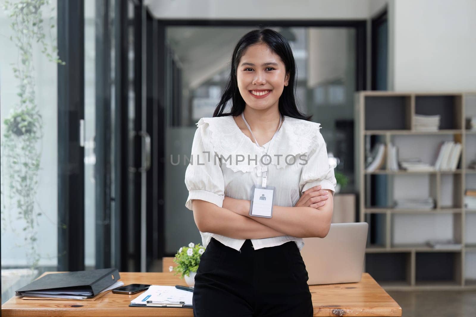 Young successful business woman entrepreneur or an office worker stands with crossed arms near a desk in a modern office, looking at the camera and smiling.
