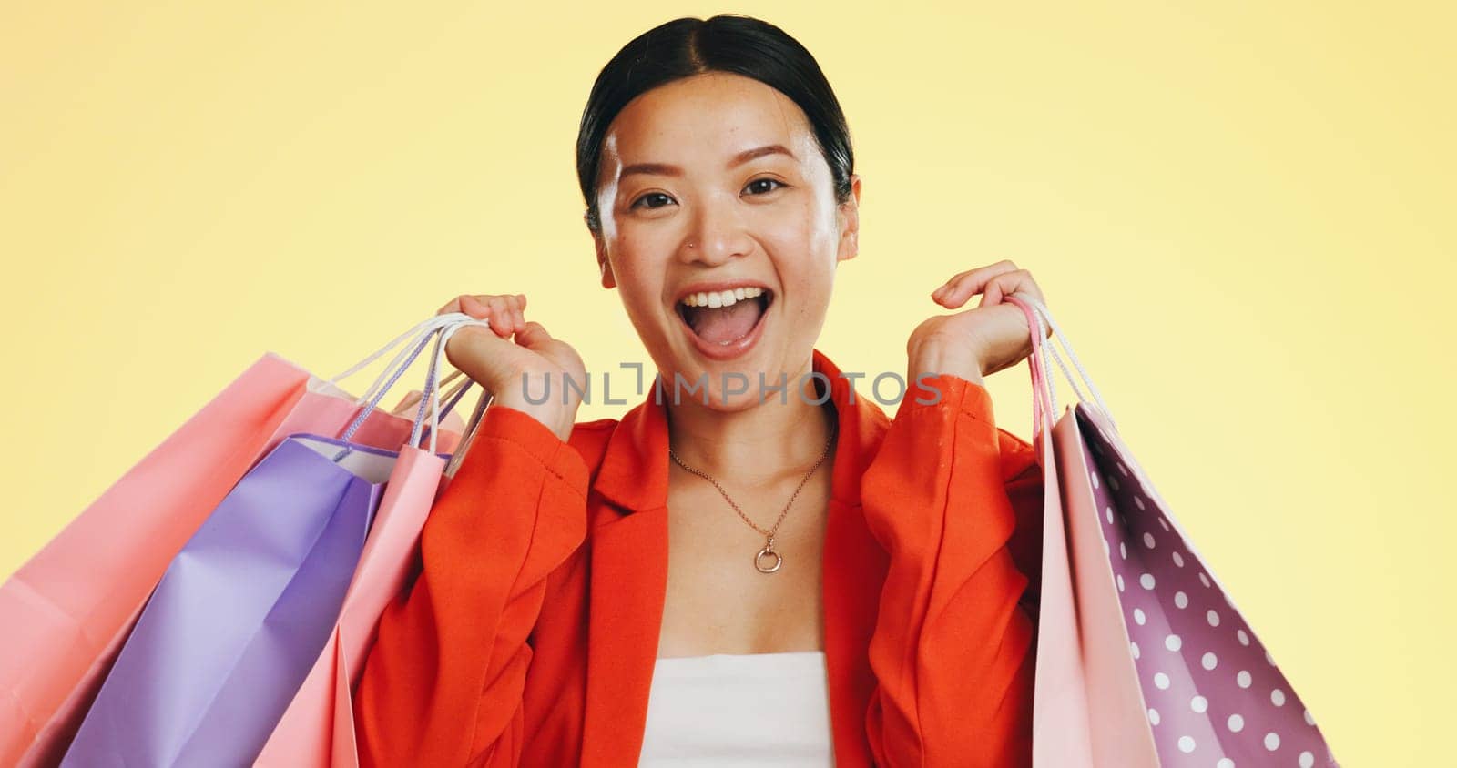 Happy, face and woman in studio with shopping, bag and boutique sale on yellow background. Portrait, excited and asian girl shopper customer with items for discount, fun and retail while isolated.