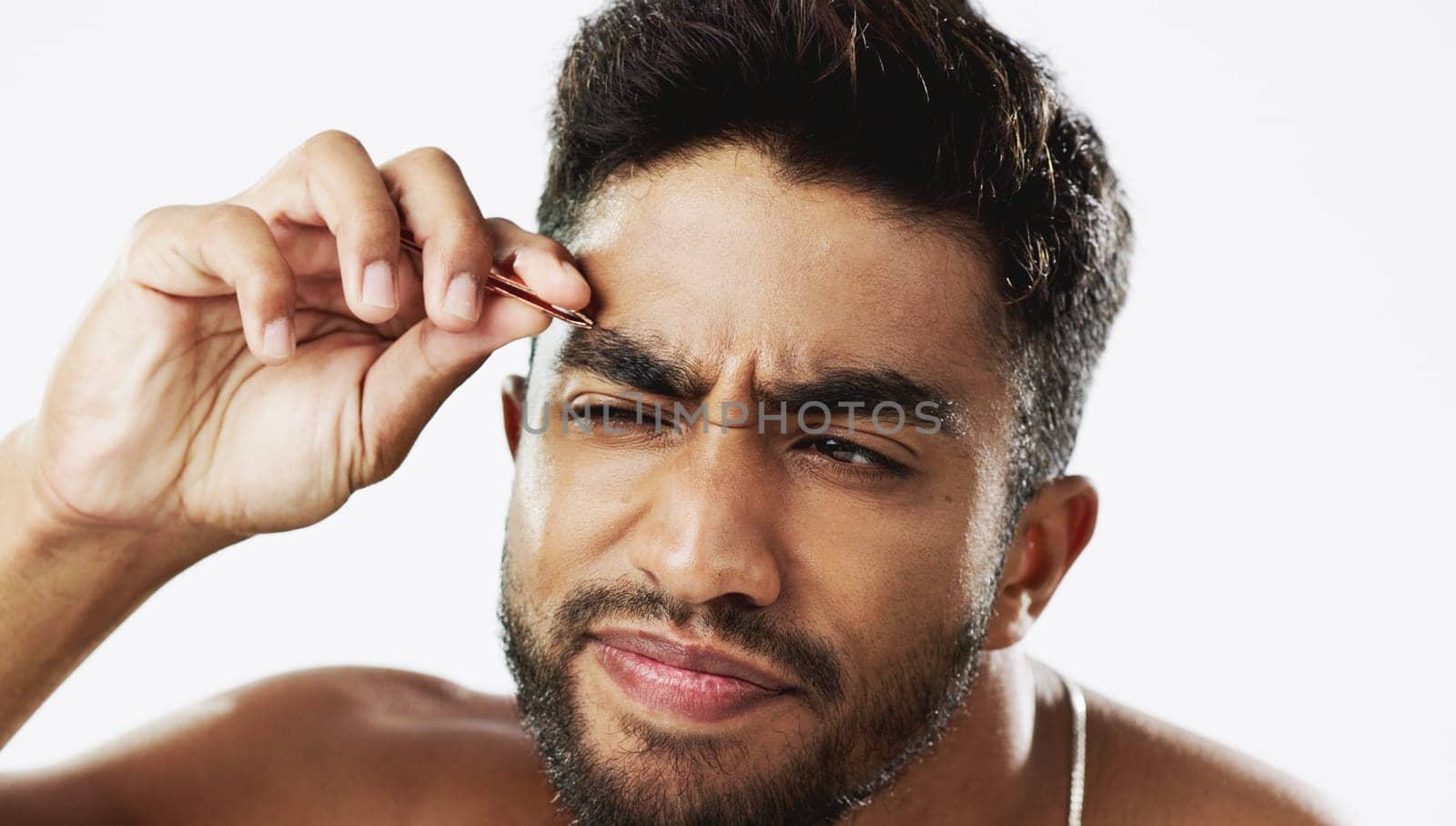 Man grooming his eyebrows with a tweezer in studio for self care, beauty and cleanliness. Hair removal, tweezing and male model from India doing facial epilation plucking routine by white background. by YuriArcurs