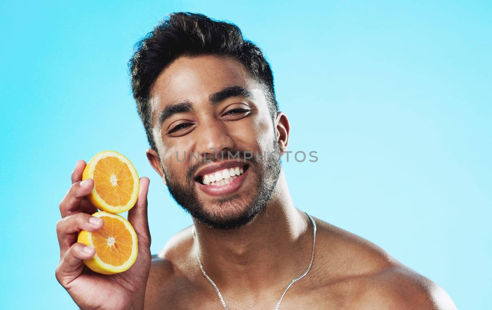 Happy, orange and face of man in studio for skincare, wellness and citrus treatment on blue background. Fruit, facial and portrait of indian male model excited for organic vitamin c skin cosmetics.