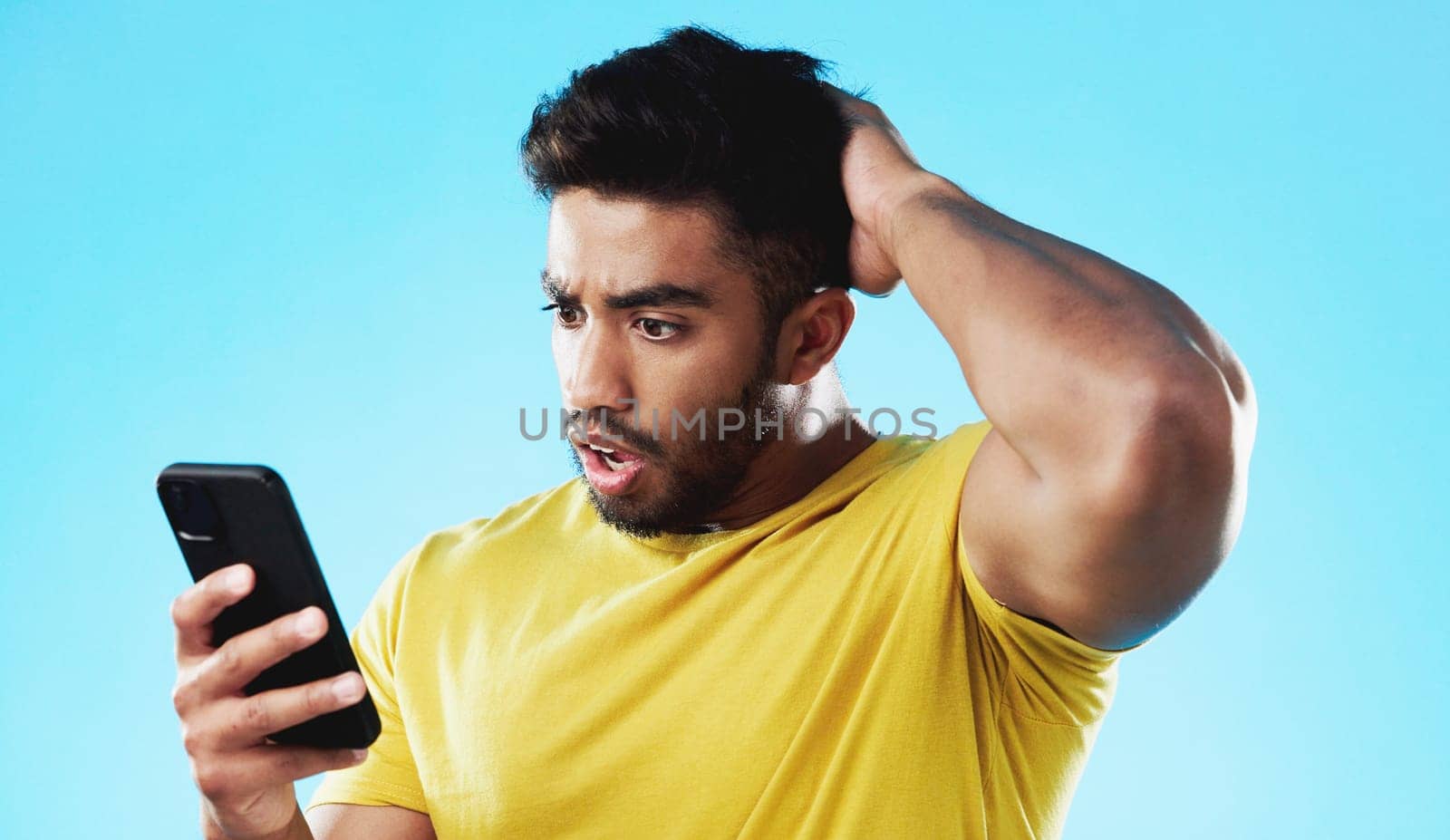 Phone, wow or bad news with a man reading a negative text message in studio on a blue background. Mobile, contact and surprise with a young male looking shocked by a social media post or announcement.