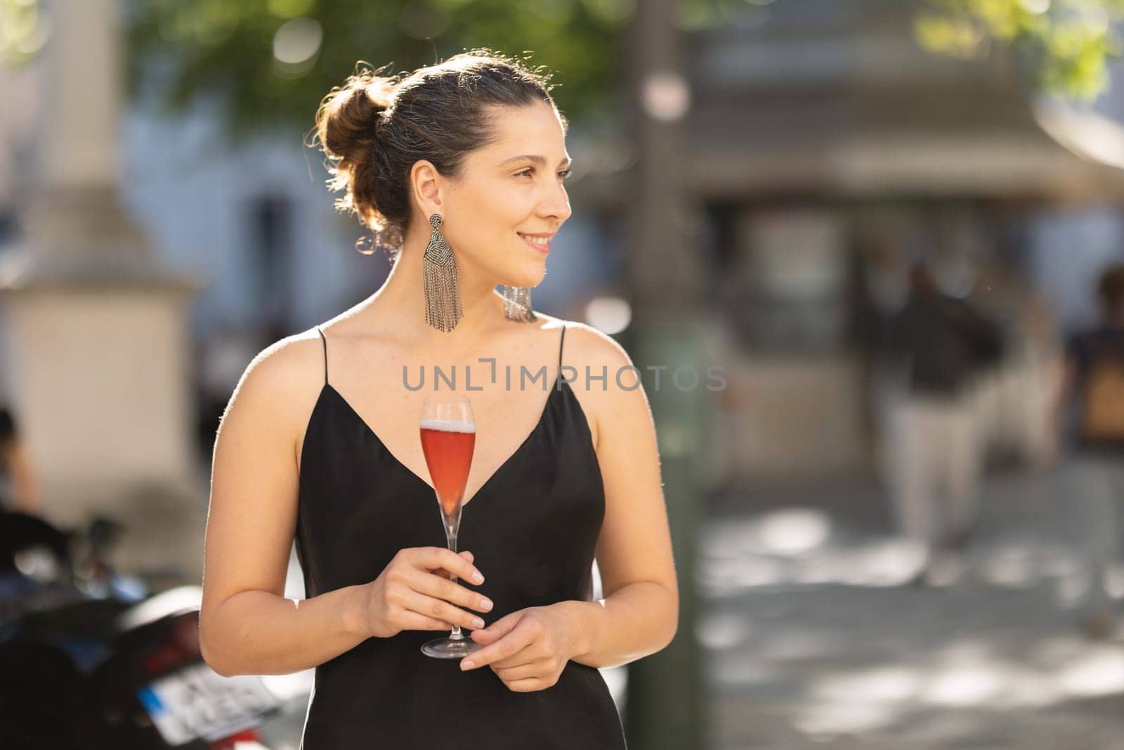 Smiling woman at casual party in elegant dress holding a drink and looking to the side. Mid shot