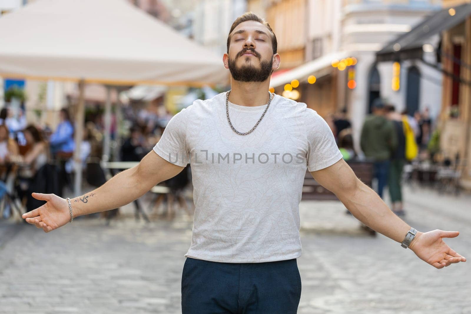 Portrait of bearded young man taking a deep breath of fresh air, relaxing, taking a break, resting, meditating outdoors. Middle eastern guy enjoying beautiful happy day in sunny urban city street