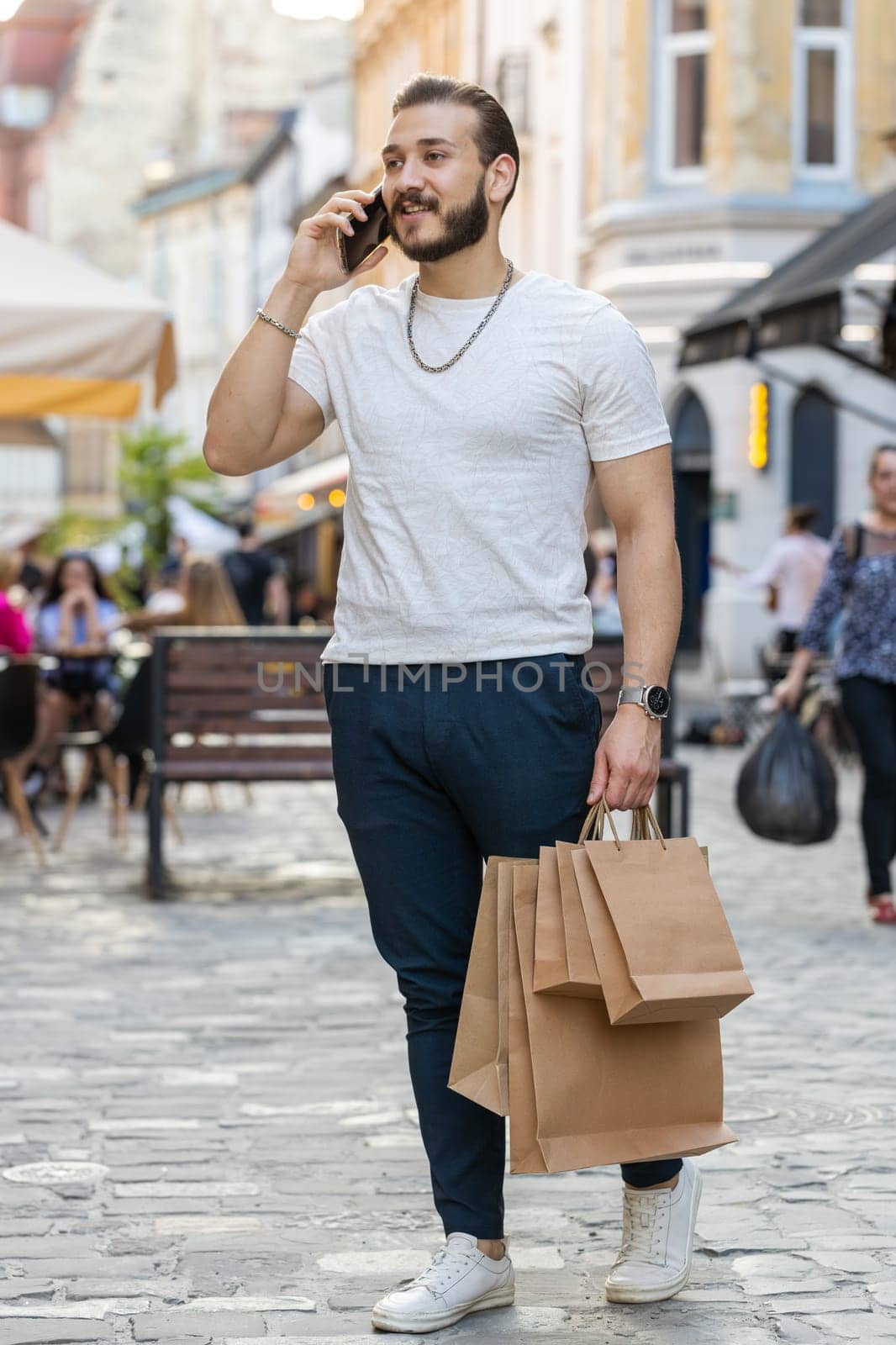 Joyful smiling bearded young man shopaholic consumer after shopping sale with full bags with gifts outdoors. Guy tourist traveler walking along the urban sunshine city street road. Town lifestyles