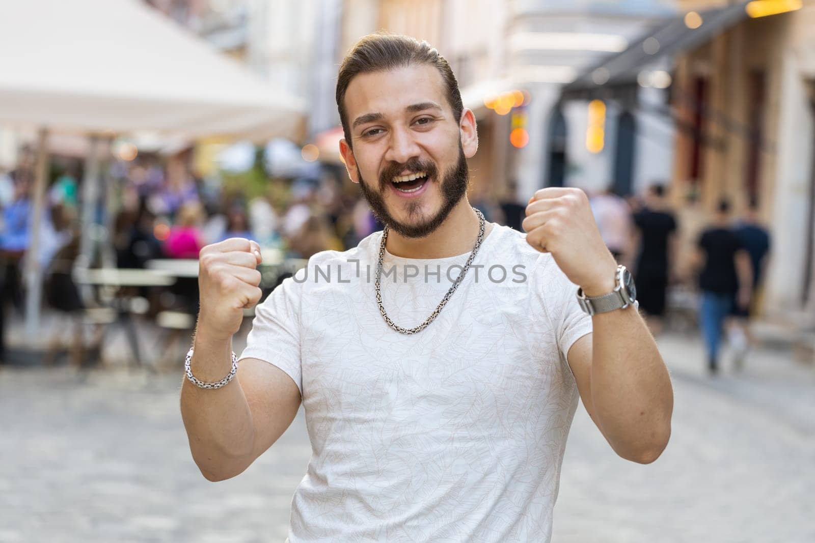 Portrait of bearded young man shouting, celebrating success, winning, goal achievement, good news, lottery jackpot luck, victory outdoors. Guy walking in urban city sunshine street. Town lifestyles