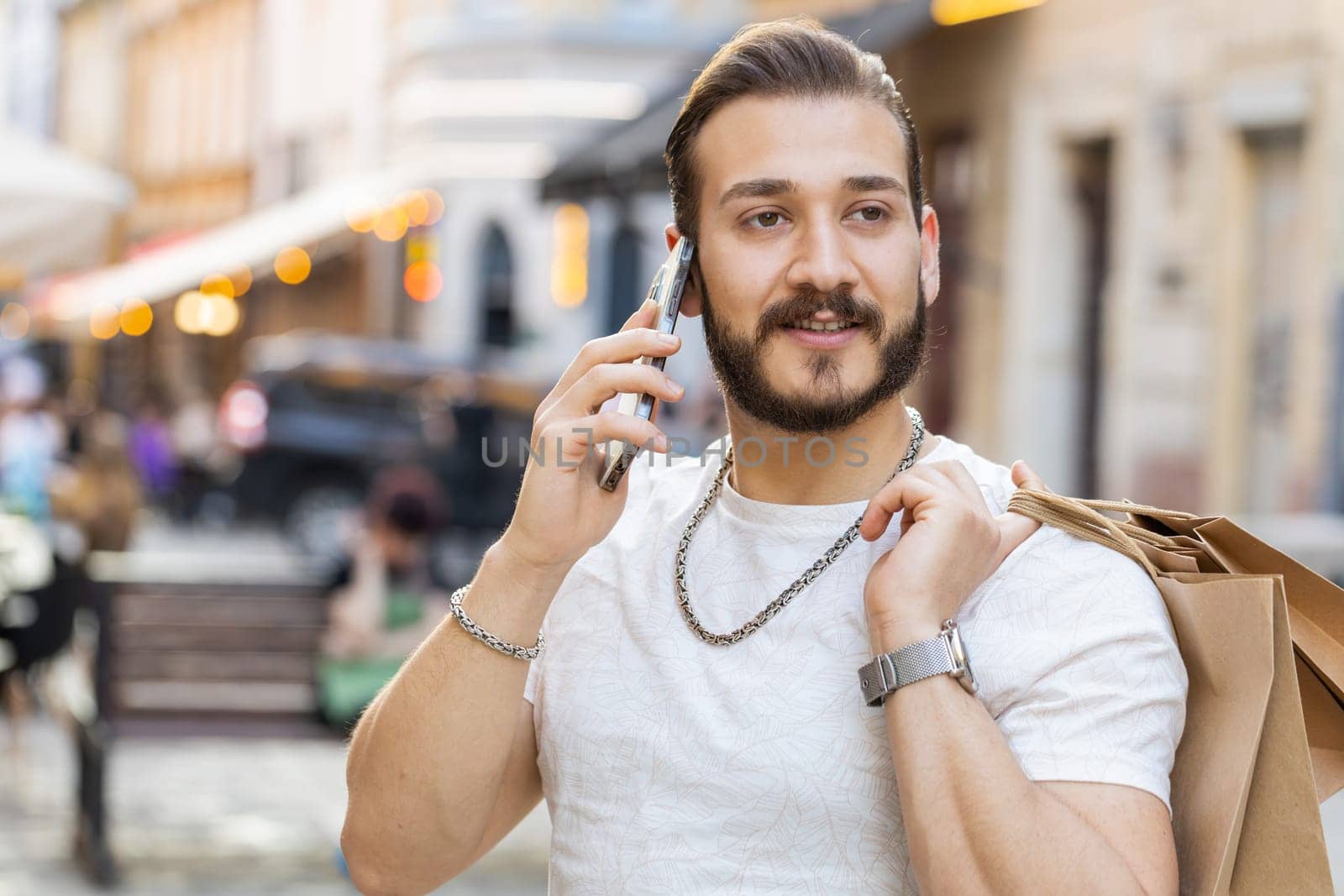 Joyful smiling bearded young man shopaholic consumer after shopping sale with full bags with gifts outdoors. Guy tourist traveler walking along the urban sunshine city street road. Town lifestyles