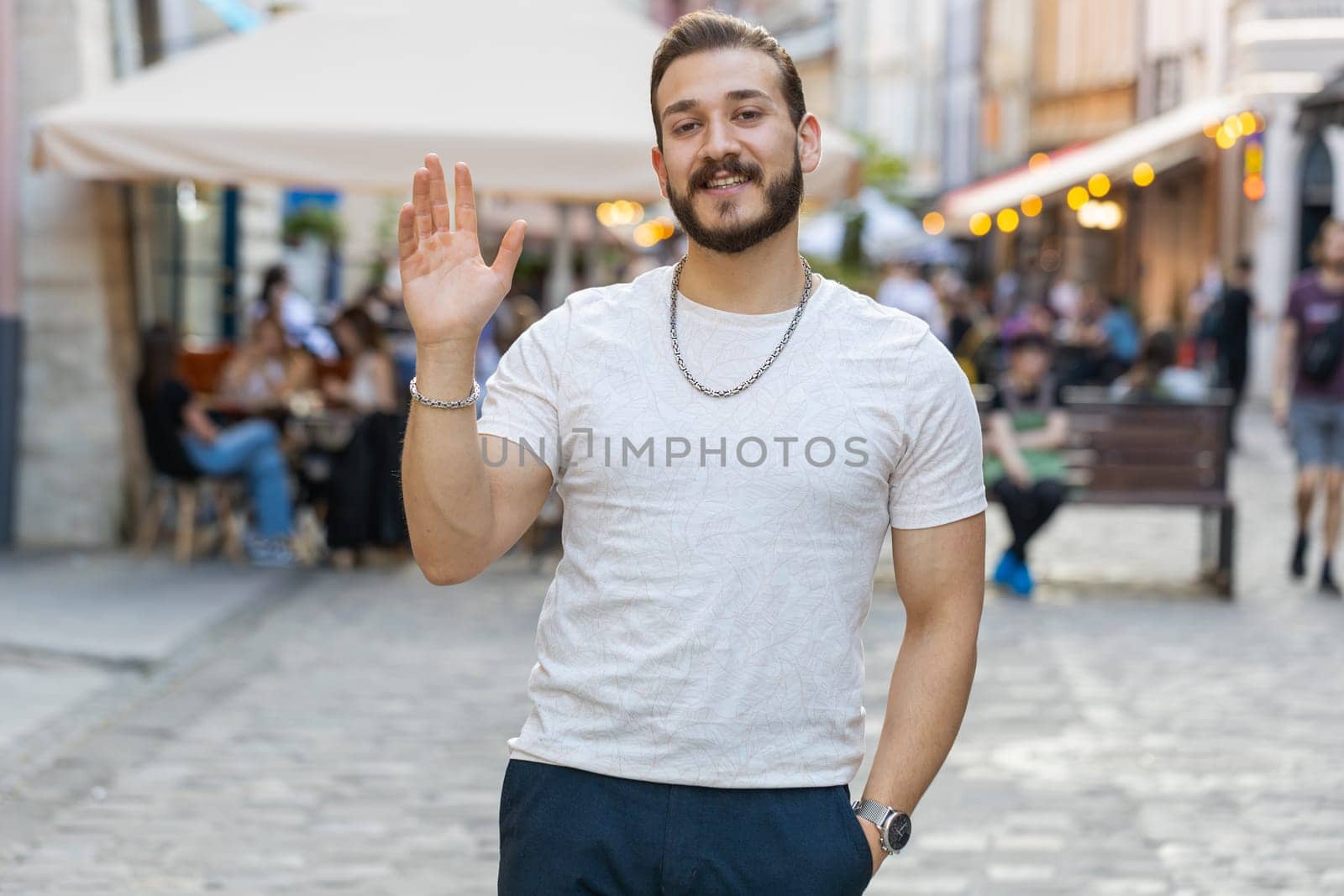 Portrait of bearded young man smiling friendly at camera, waving hands gesturing hello, hi, greeting or goodbye, welcoming with hospitable expression outdoors. Guy walking in urban city town street