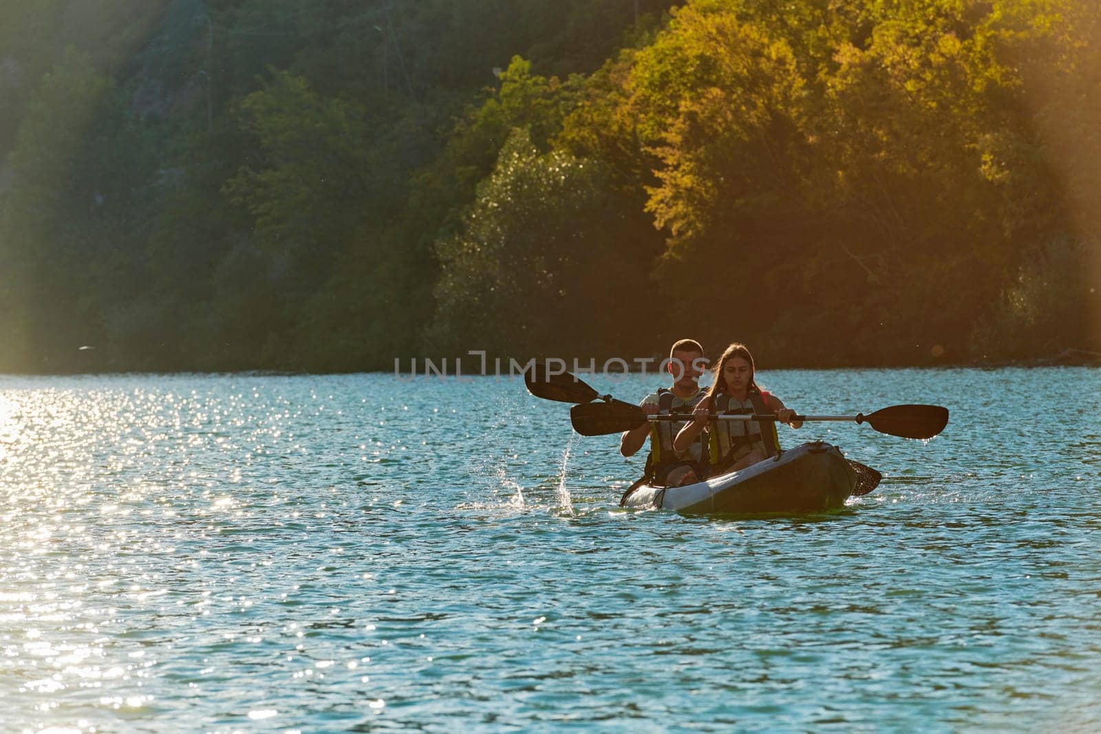 A young couple enjoying an idyllic kayak ride in the middle of a beautiful river surrounded by forest greenery in sunset time.