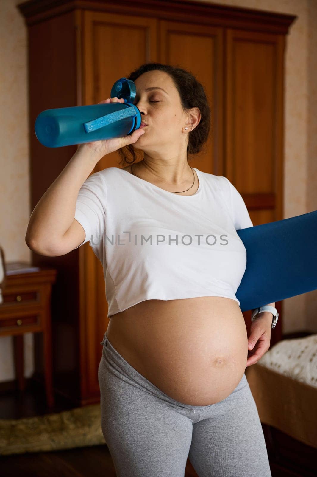 Pregnant sporty woman drinks water, renewing aqua balance after fitness workout, holding an exercise mat, standing at home interior, enjoying active healthy lifestyle in pregnancy and maternity leave