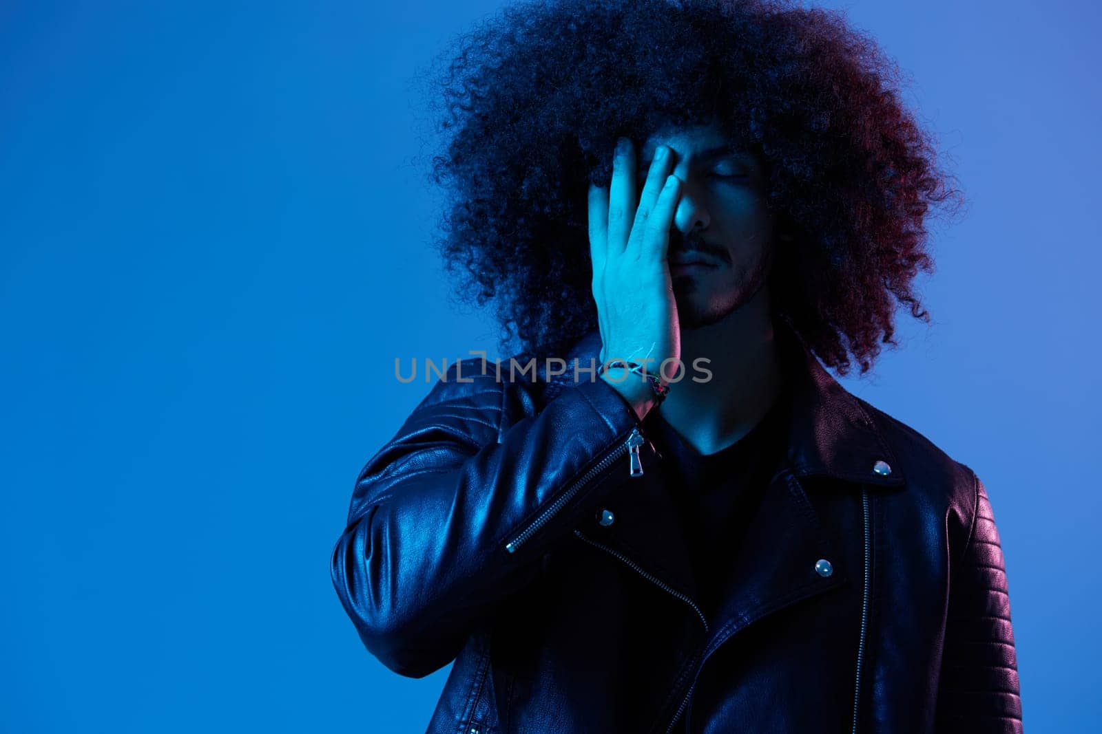 Portrait of fashion man with curly hair on blue background multinational, colored light, black leather jacket trend, modern concept. High quality photo