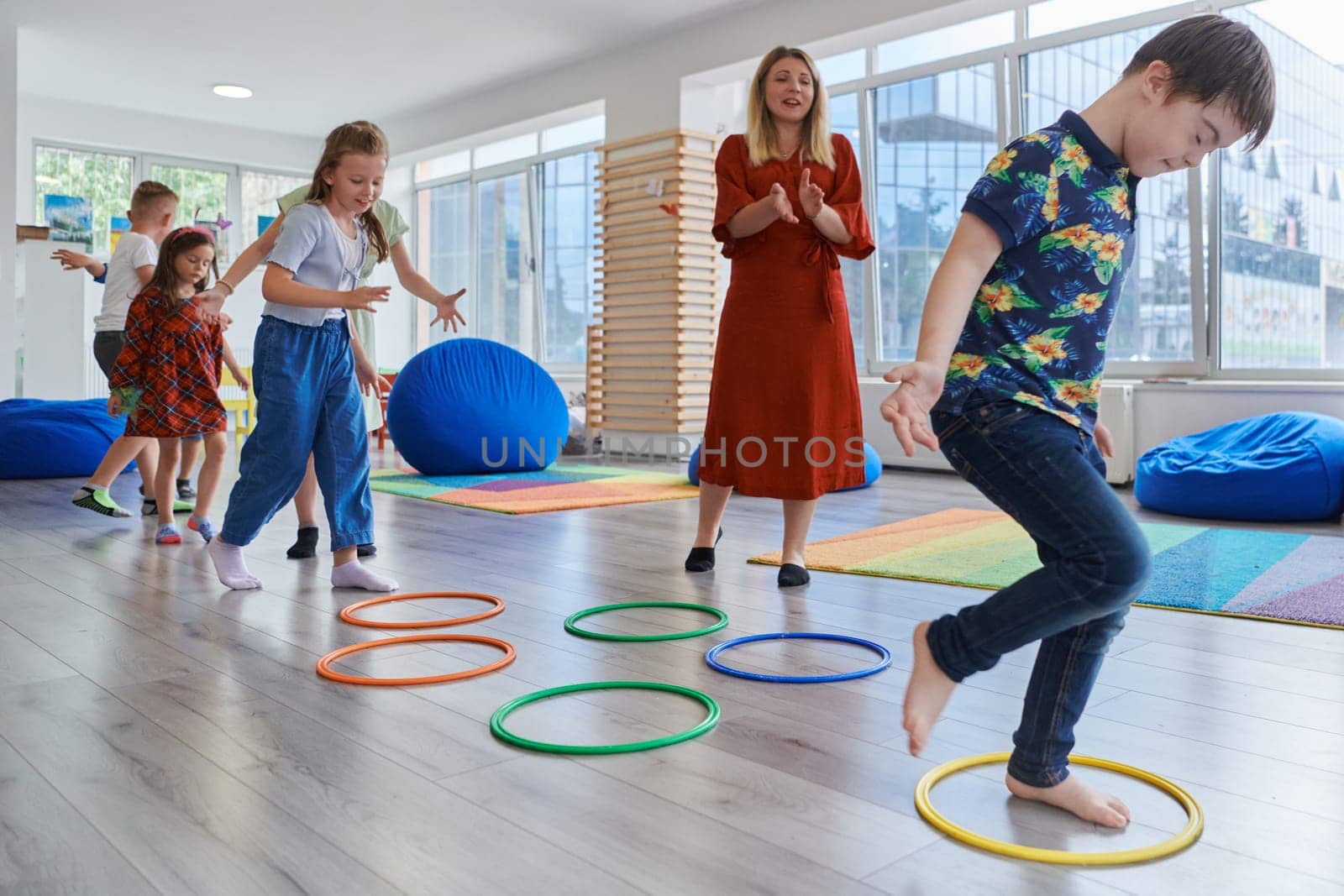 Small nursery school children with female teacher on floor indoors in classroom, doing exercise. Jumping over hula hoop circles track on the floor