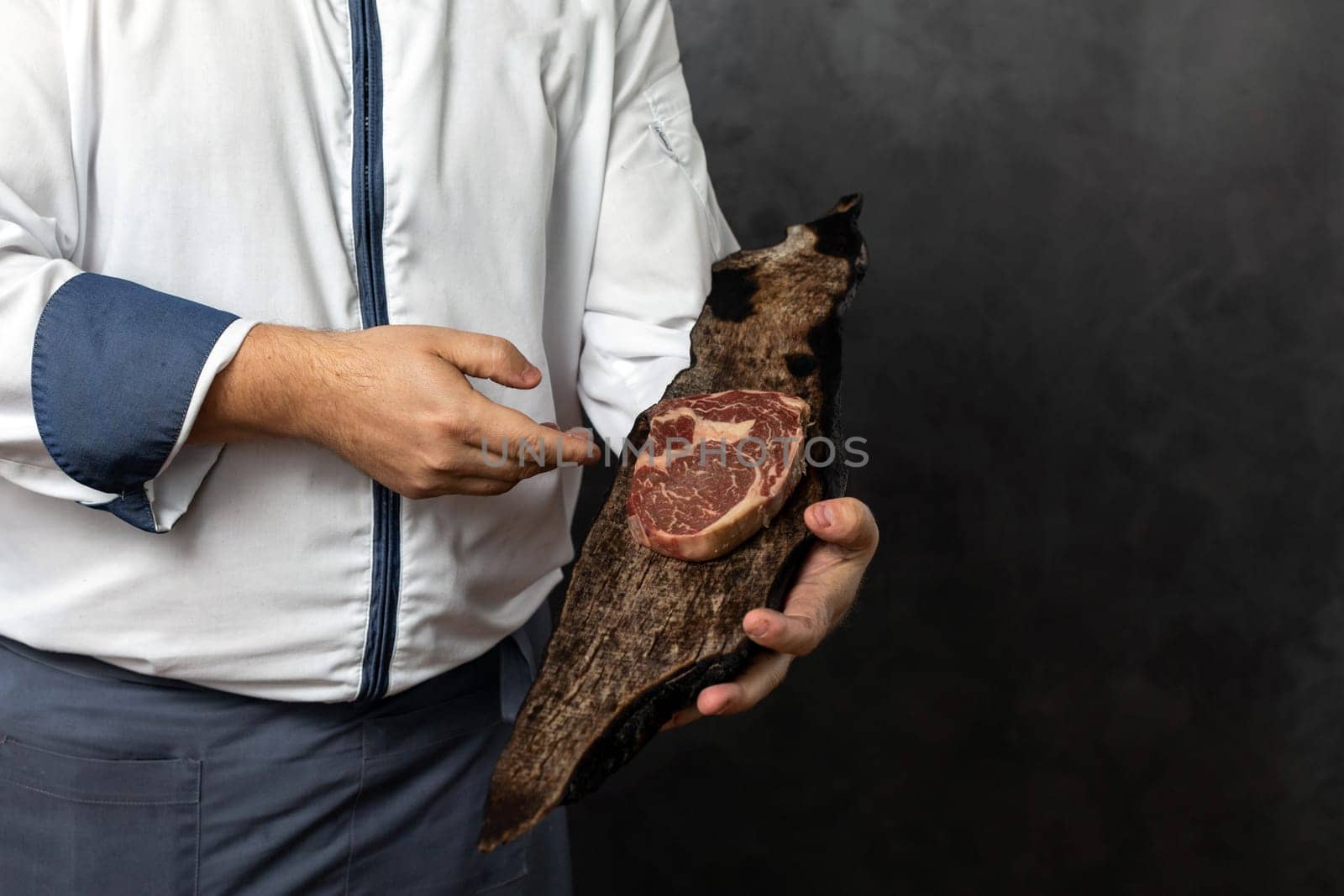 Crop anonymous male in uniform holding piece of fresh delicious meat on wooden board against grunge black background