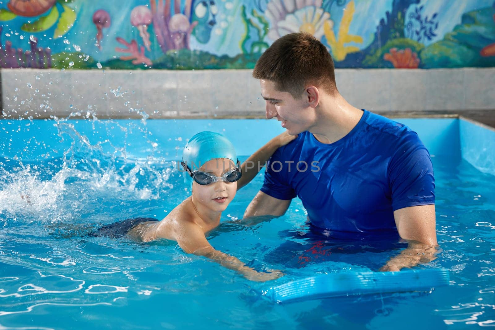 Little boy learning to swim in indoor pool with pool board. Swimming lesson. Active child swims in water with teacher