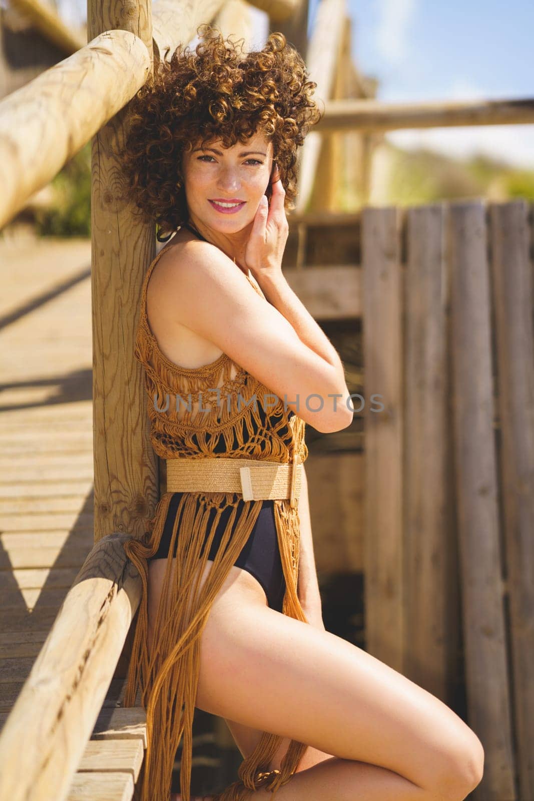 Young happy curly haired female in swimsuit, and brown knitted beach cover up leaning on wooden railing touching face gently while standing on coast looking at camera