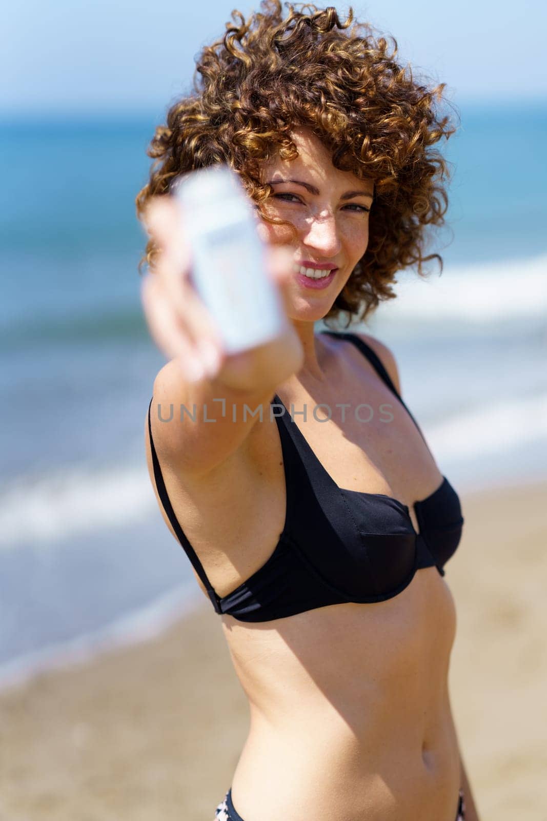Smiling young female in bikini standing on sandy beach near waving sea and showing white bottle of sunblock cream