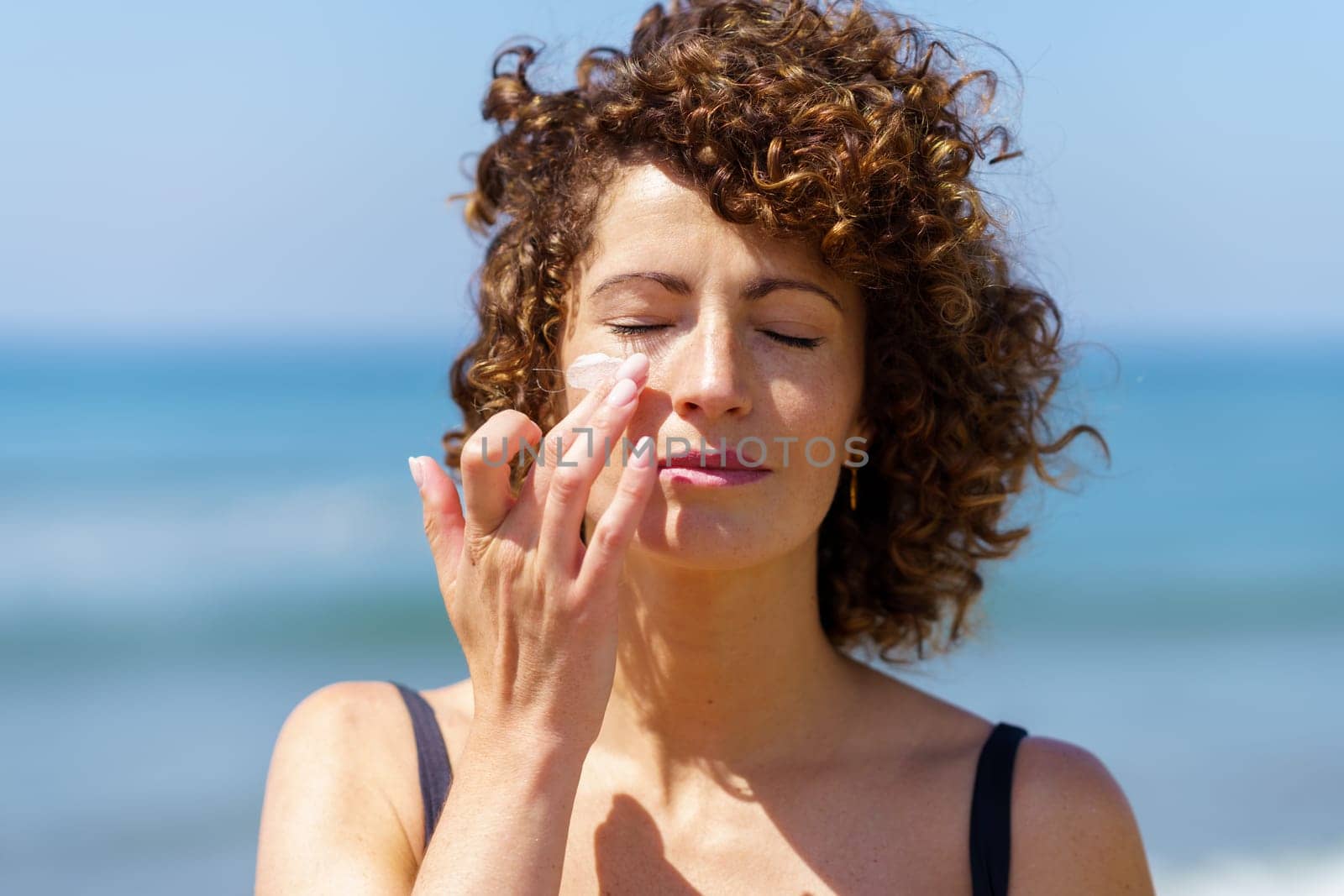 Young female with curly hair applying sunblock cream on face while standing against blurred sea on sunny day