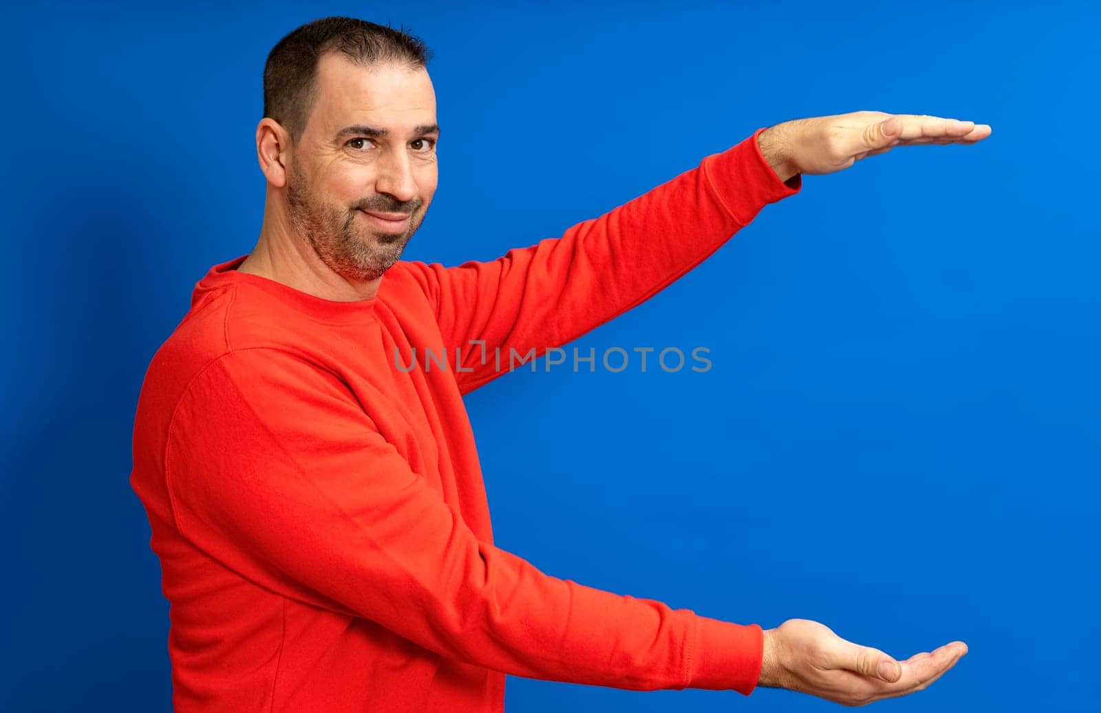Bearded hispanic man wearing a red sweater with open arms in profile offering something while smiling at camera, isolated over blue background