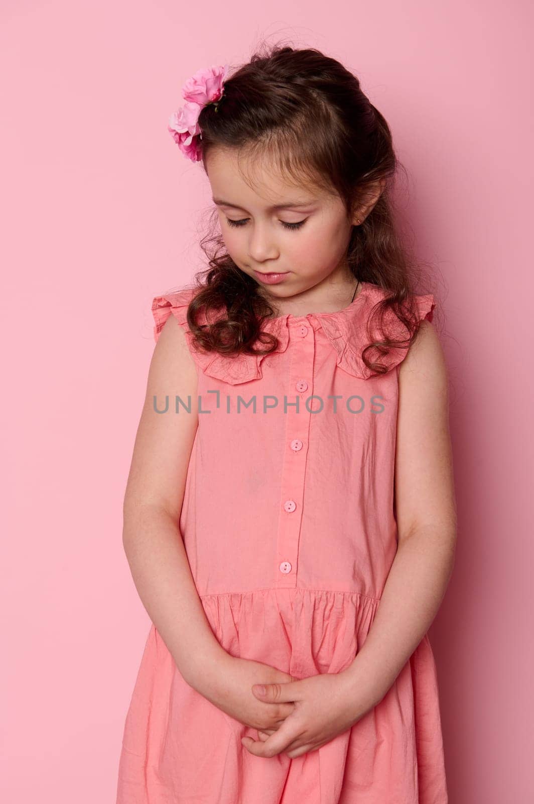 Beautiful coquette, shy child, adorable little girl 5-6 years old, standing over pink isolated background. Happy carefree childhood concept. Portrait of a preschooler child. Kids fashions and beauty