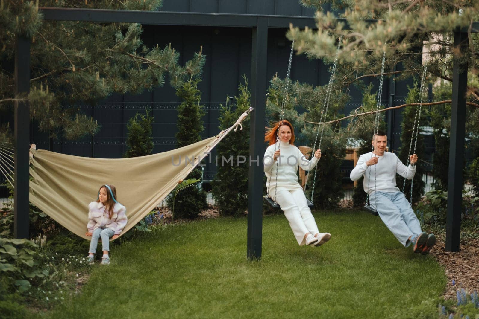 Mom and dad are riding on a swing, and their daughter is on a hammock next to them. The family is resting on a swing.