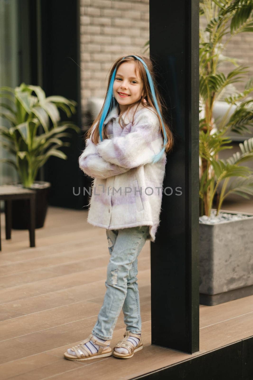 A little girl in a jacket with long hair is standing on the terrace at the house.