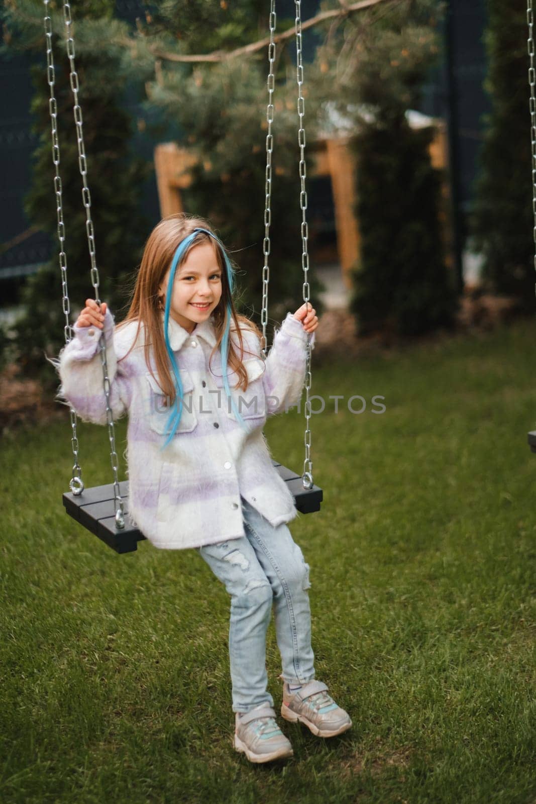 A beautiful girl with long hair in a jacket rides on a swing in the evening on the street by Lobachad