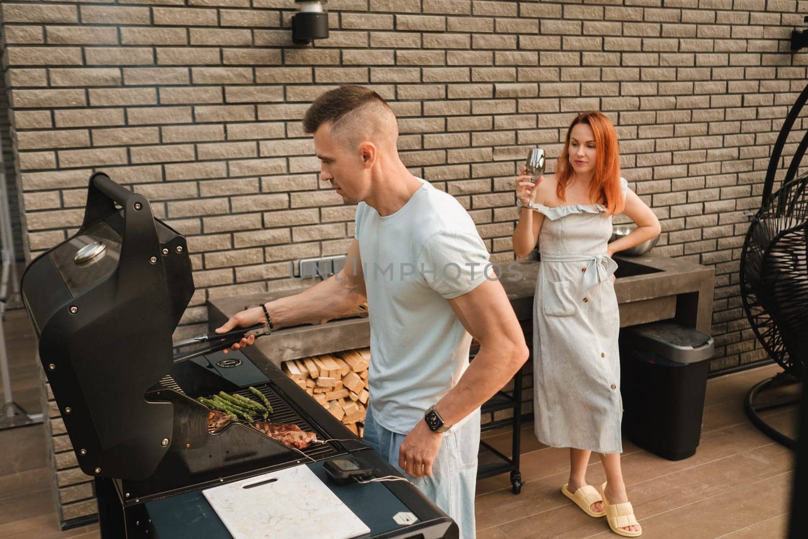 A married couple cooks grilled meat together on their terrace by Lobachad
