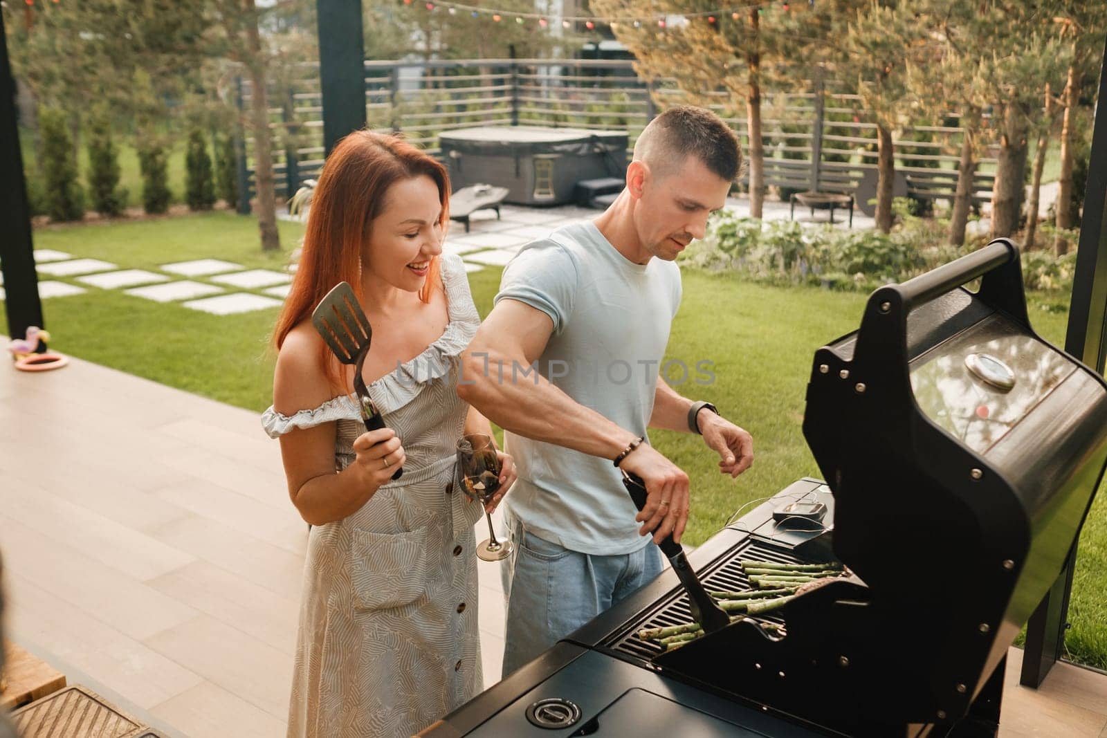 A married couple cooks grilled meat together on their terrace.