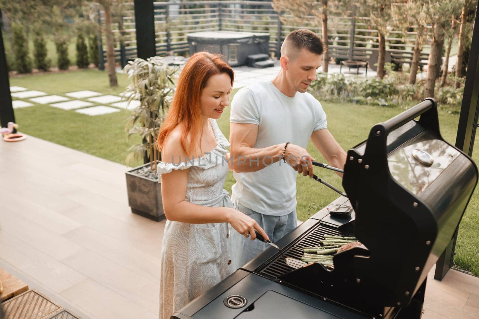 A married couple cooks grilled meat together on their terrace.