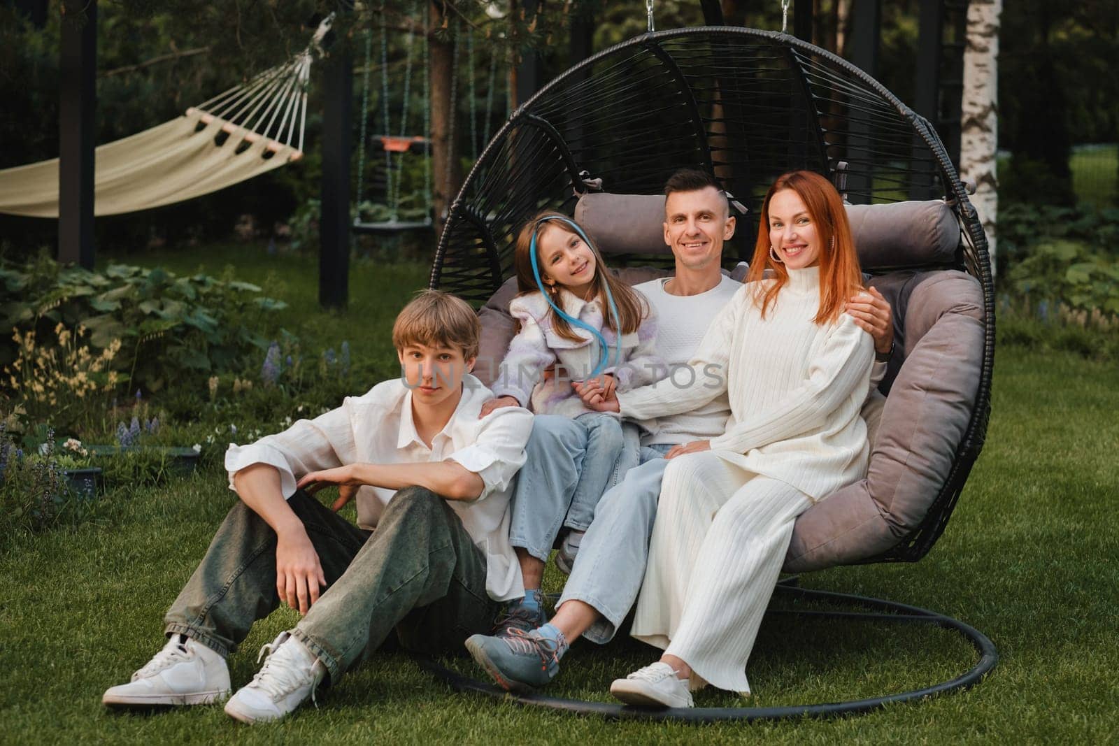 A happy family is sitting in a hammock on the lawn near the house by Lobachad