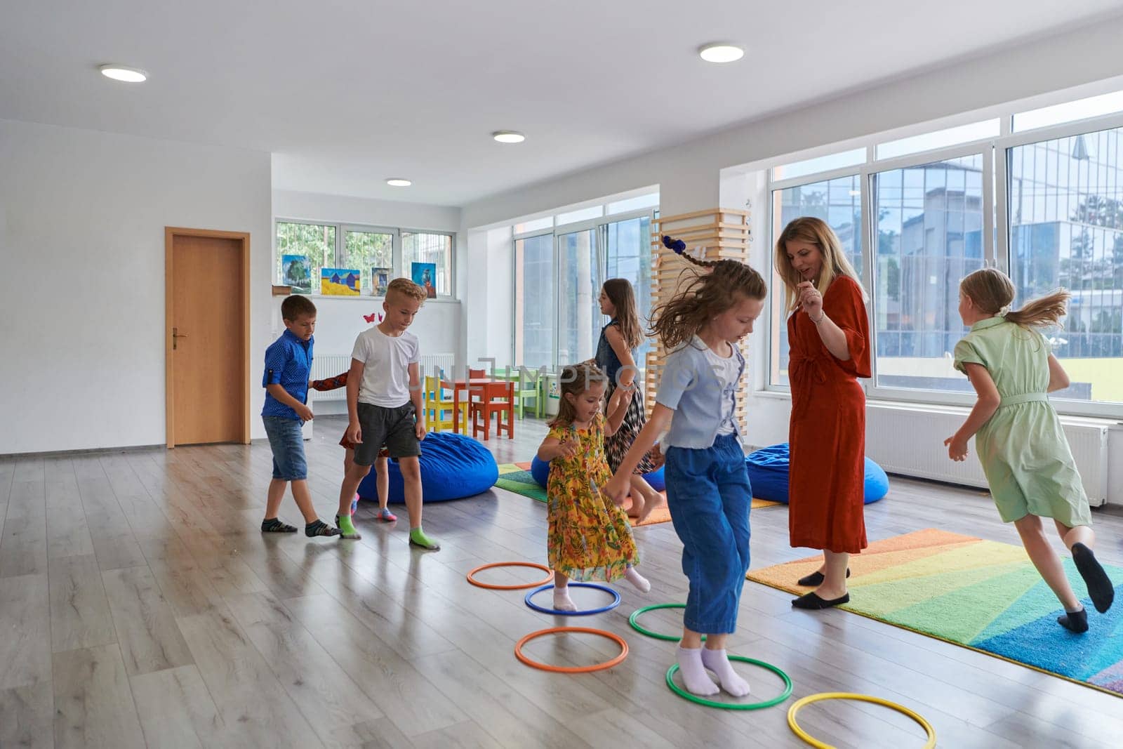 Small nursery school children with female teacher on floor indoors in classroom, doing exercise. Jumping over hula hoop circles track on the floor