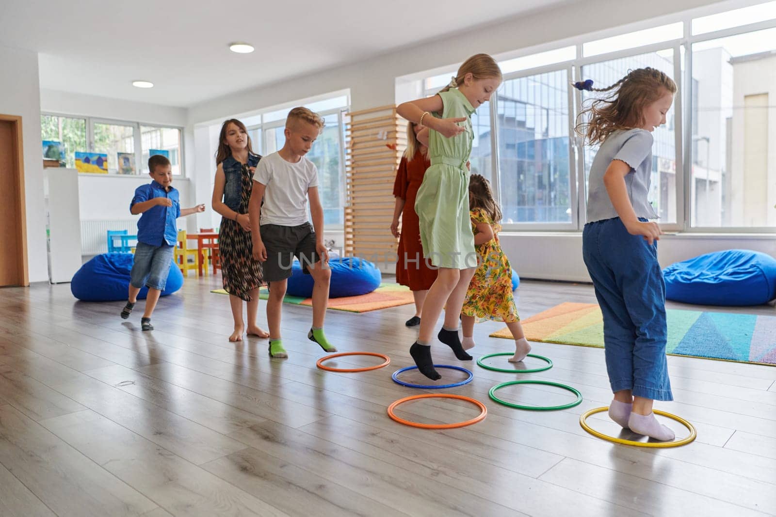 Small nursery school children with female teacher on floor indoors in classroom, doing exercise. Jumping over hula hoop circles track on the floor. by dotshock