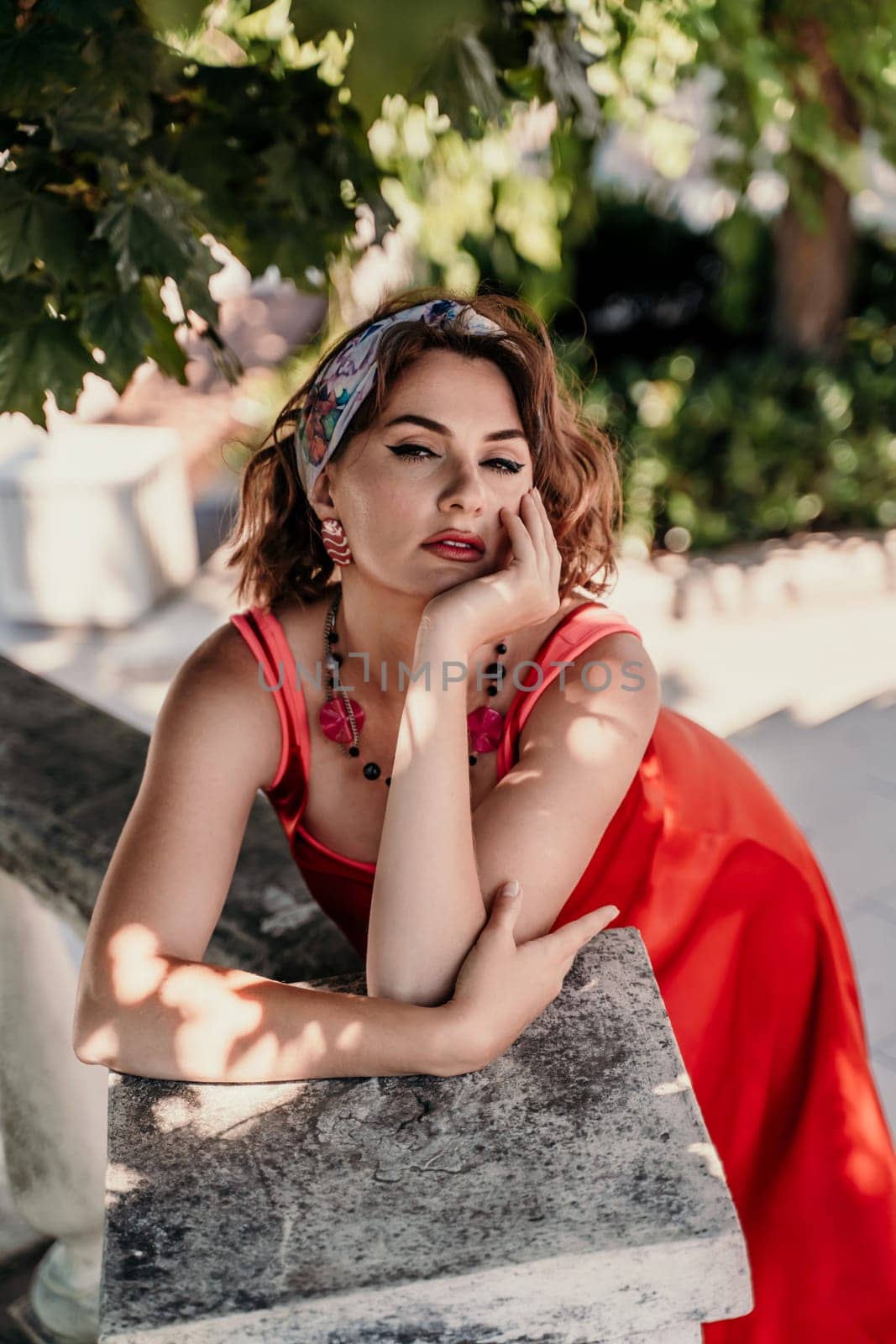 A pretty woman in a red silk dress and a bandage on her head smiles against the background of the leaves of a tree. She is leaning on the coop and looking into the camera. Vertical photo. by Matiunina