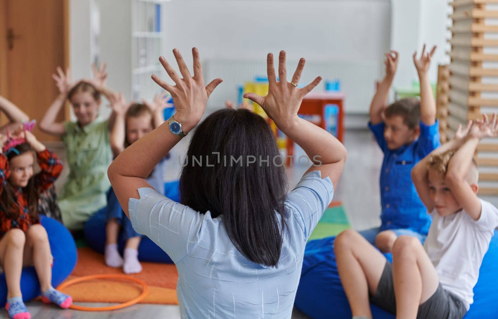 A happy female teacher sitting and playing hand games with a group of little schoolchildren by dotshock