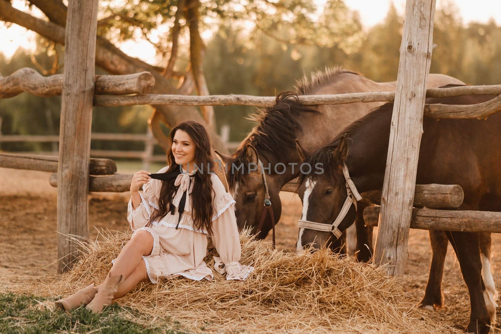 a pregnant woman in a dress in the countryside is sitting on the hay near the horses.