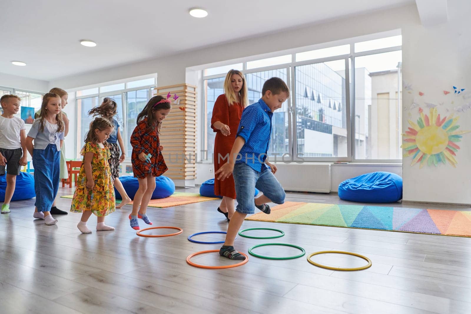 Small nursery school children with female teacher on floor indoors in classroom, doing exercise. Jumping over hula hoop circles track on the floor. by dotshock