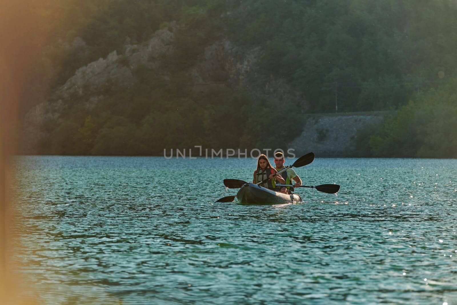 A young couple enjoying an idyllic kayak ride in the middle of a beautiful river surrounded by forest greenery in sunset time.