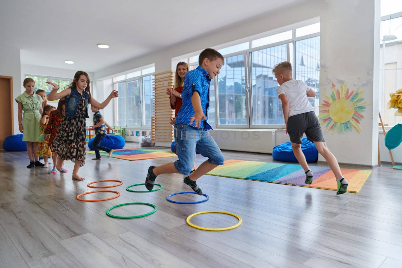 Small nursery school children with female teacher on floor indoors in classroom, doing exercise. Jumping over hula hoop circles track on the floor