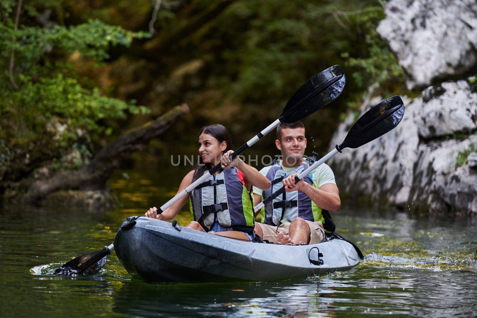 A young couple enjoying an idyllic kayak ride in the middle of a beautiful river surrounded by forest greenery by dotshock