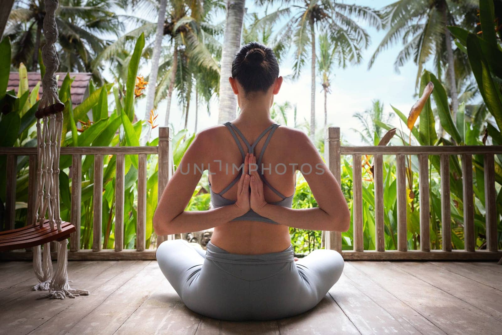 Back view of young woman doing reverse namaste mudra while meditating in tropical vacation resort. Reverse prayer pose. Yoga and spirituality concepts.