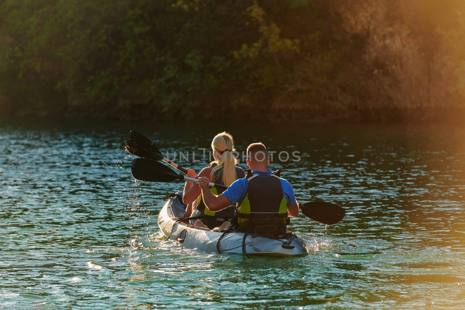 A young couple enjoying an idyllic kayak ride in the middle of a beautiful river surrounded by forest greenery in sunset time.