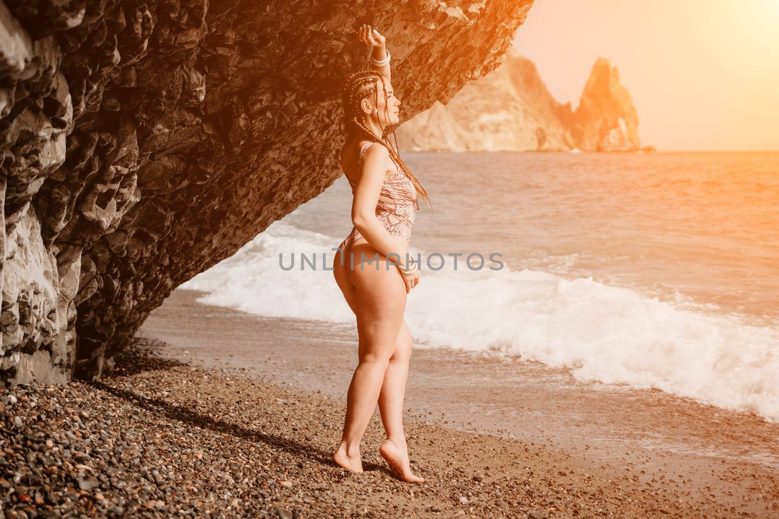 A tourist woman in bikini on beach enjoys the turquoise sea during her summer holiday. Beach vacation. Woman with braids dreadlocks standing with her arms raised near basalt rock enjoying beach ocean by panophotograph