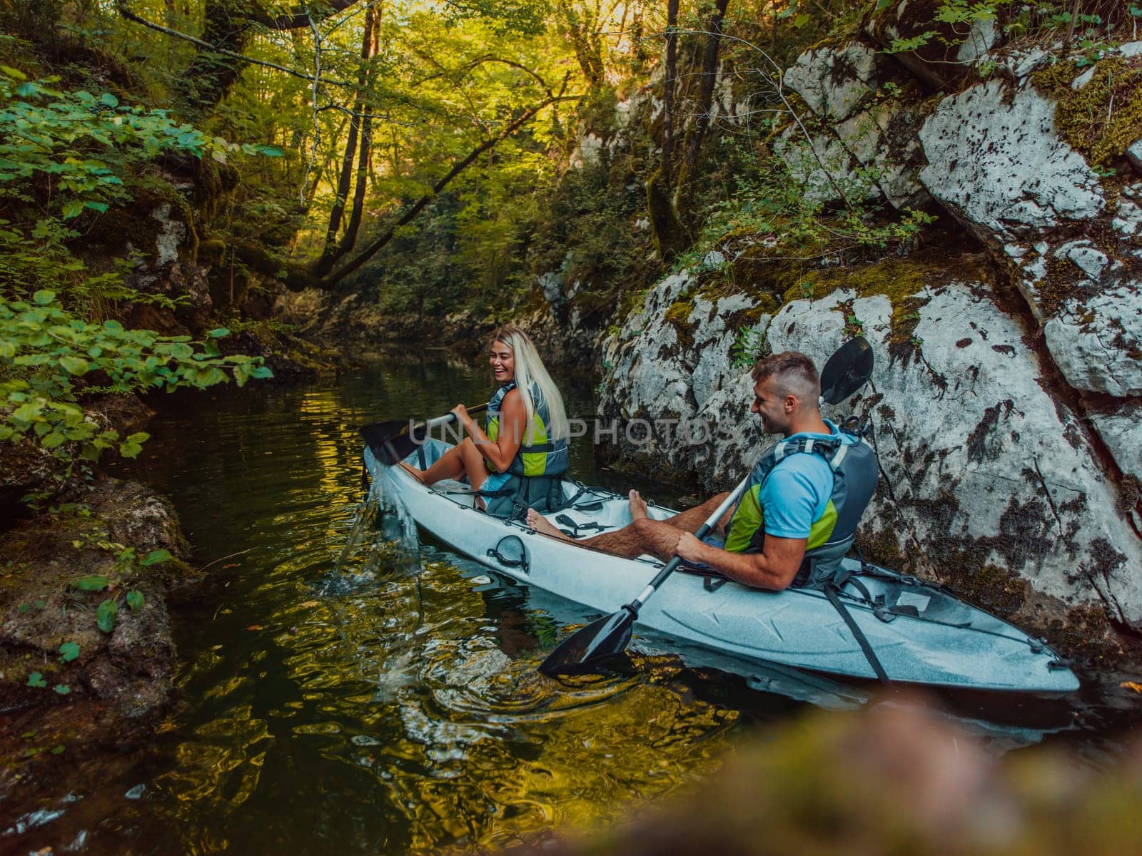 A young couple enjoying an idyllic kayak ride in the middle of a beautiful river surrounded by forest greenery by dotshock