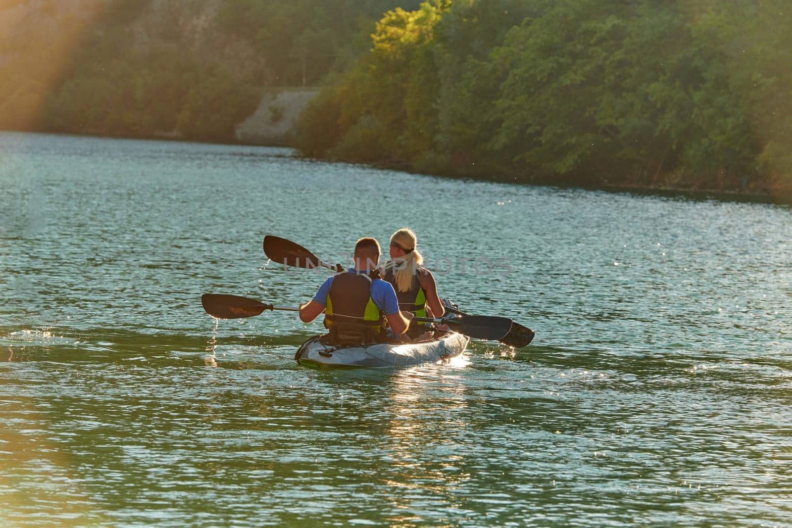 A young couple enjoying an idyllic kayak ride in the middle of a beautiful river surrounded by forest greenery in sunset time.