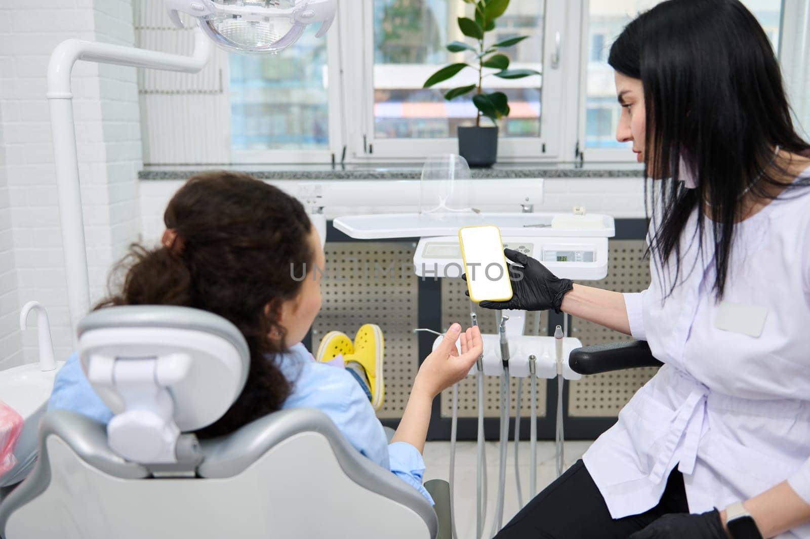 Rear view woman dentist holding smartphone with white blank screen and patient at dental appointment in dentistry clinic by artgf