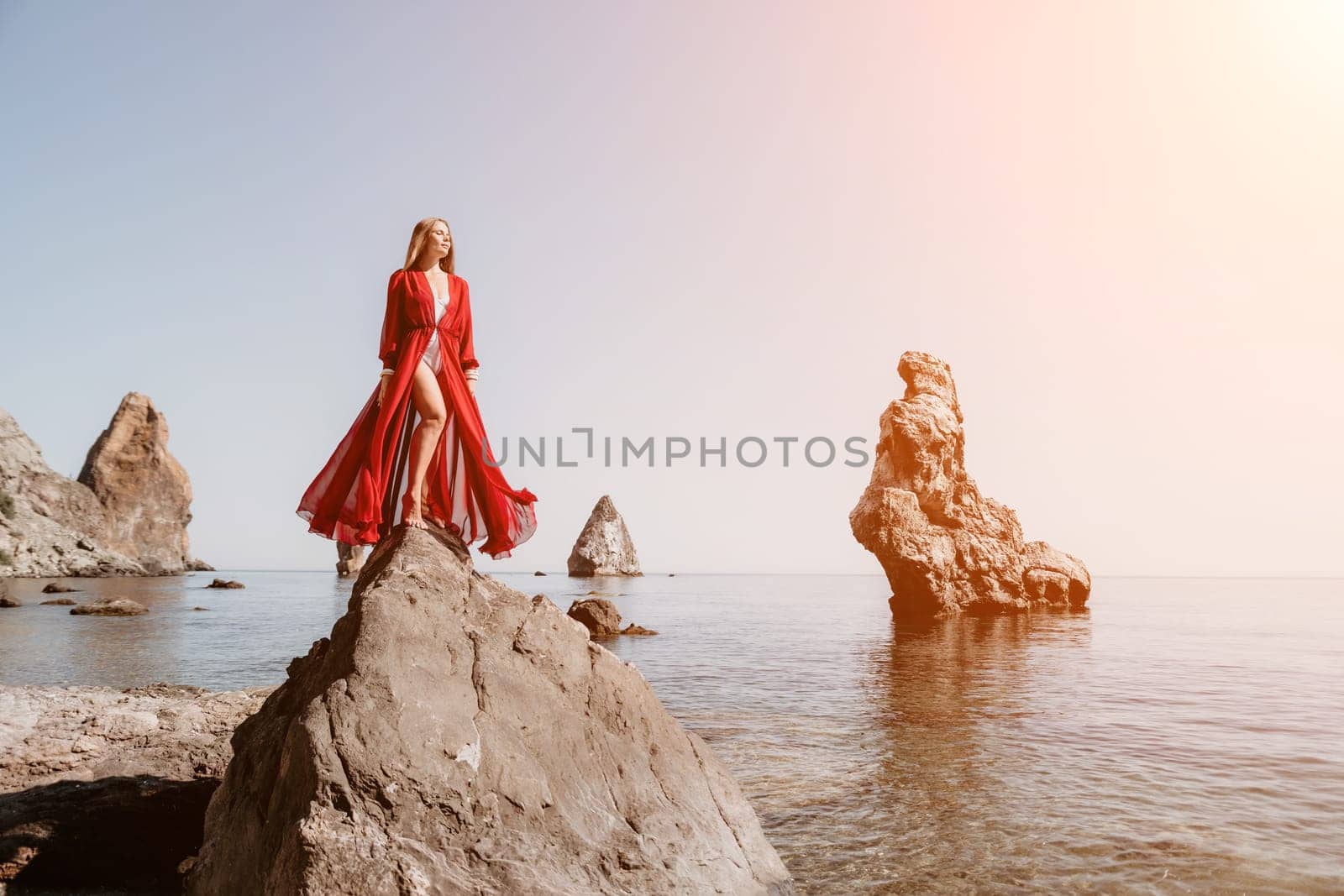 Woman travel sea. Young Happy woman in a long red dress posing on a beach near the sea on background of volcanic rocks, like in Iceland, sharing travel adventure journey by panophotograph