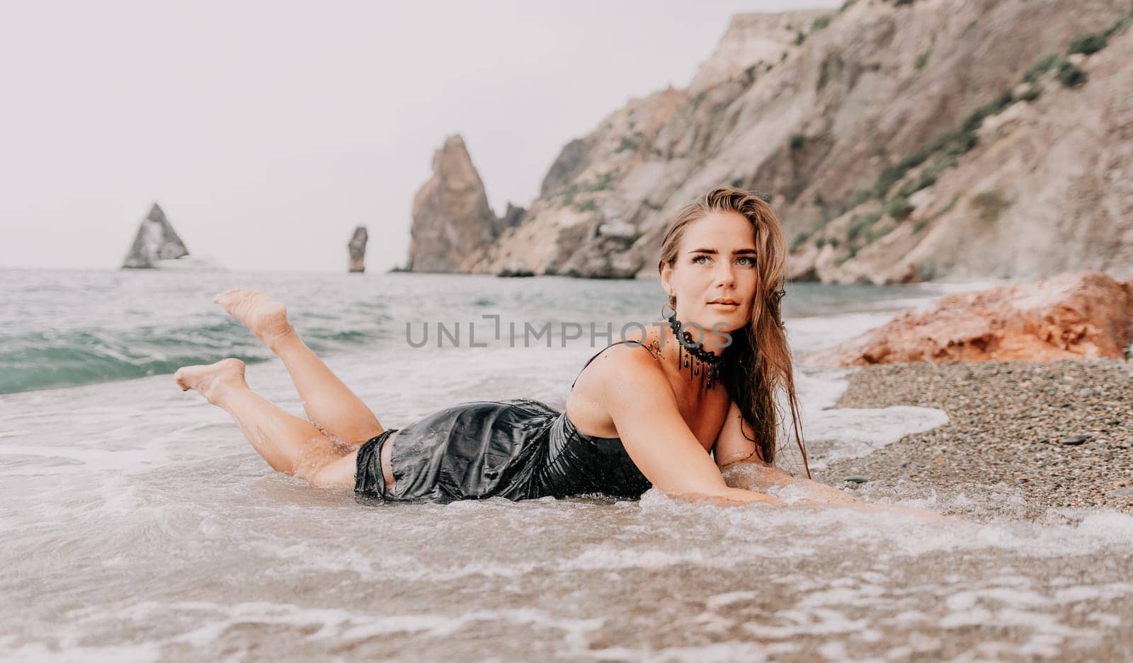 Woman summer travel sea. Happy tourist in black dress enjoy taking picture outdoors for memories. Woman traveler posing on sea beach surrounded by volcanic mountains, sharing travel adventure journey by panophotograph