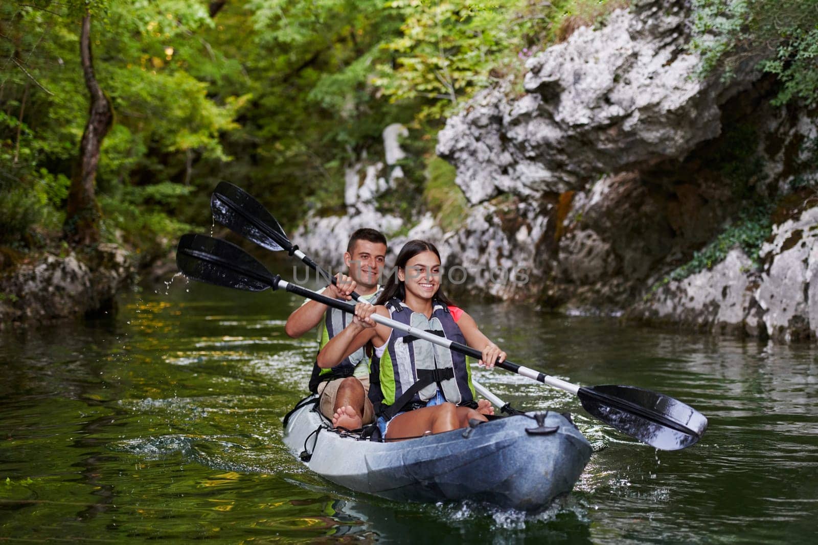 A young couple enjoying an idyllic kayak ride in the middle of a beautiful river surrounded by forest greenery.