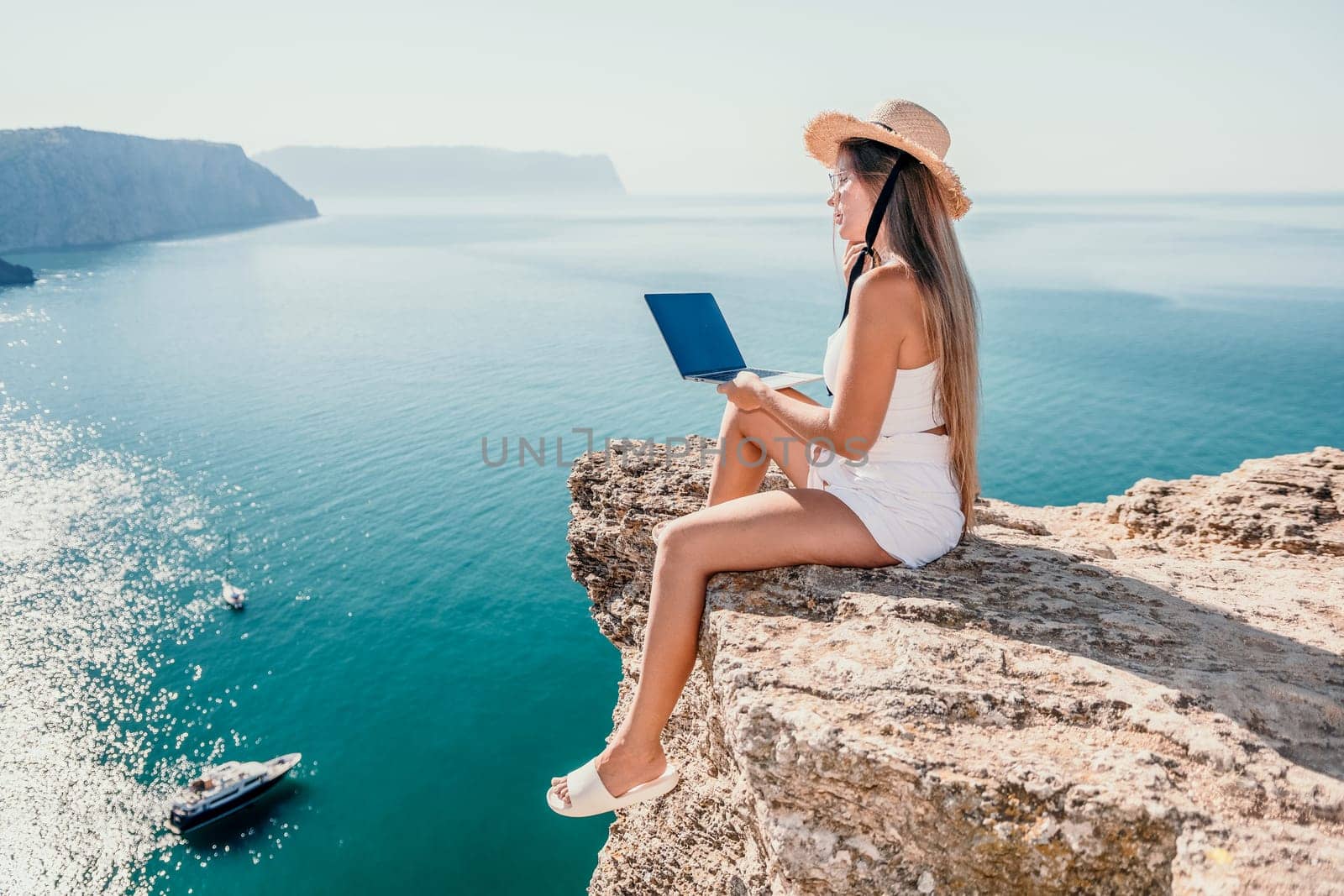 Successful business woman in yellow hat working on laptop by the sea. Pretty lady typing on computer at summer day outdoors. Freelance, travel and holidays concept.