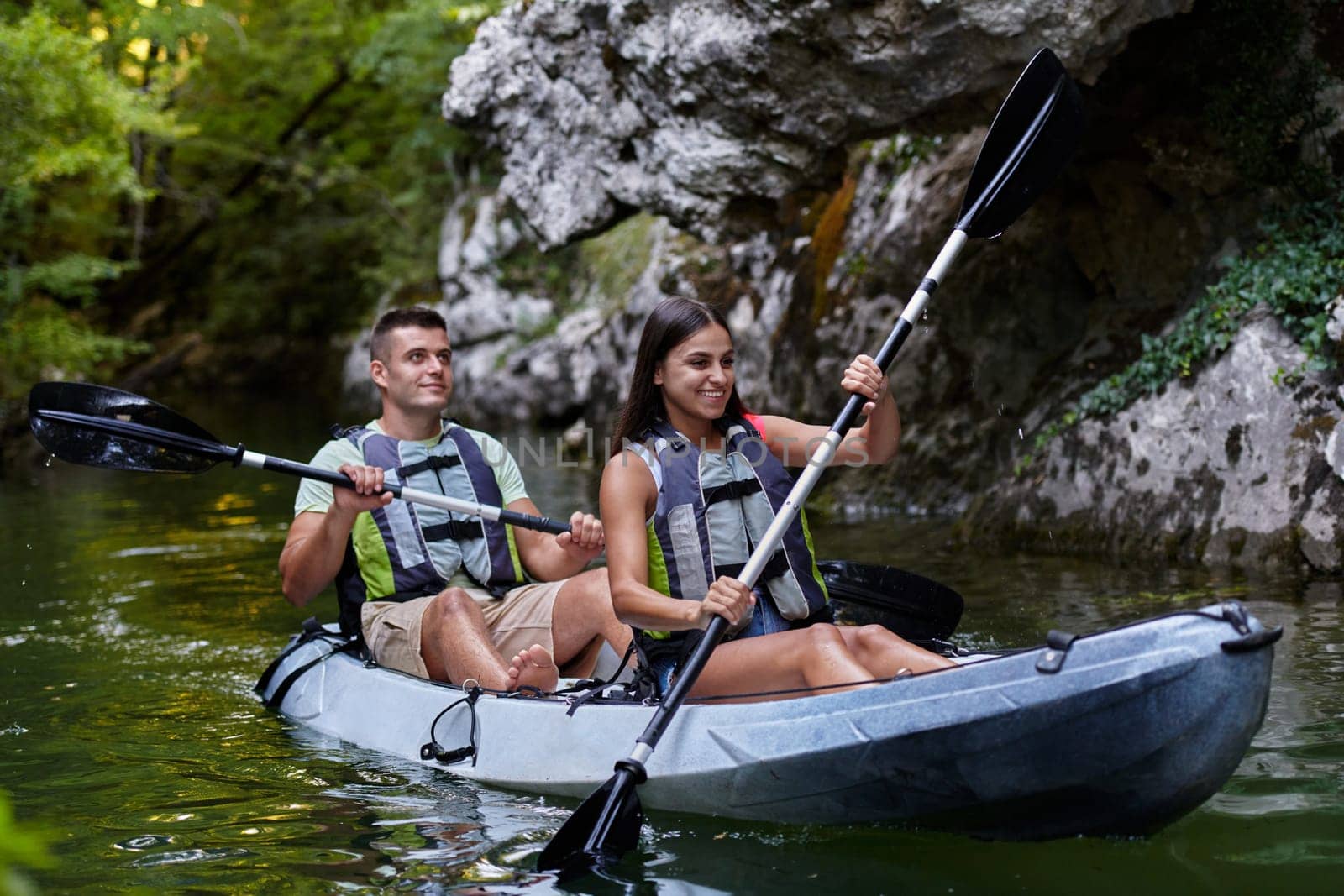 A young couple enjoying an idyllic kayak ride in the middle of a beautiful river surrounded by forest greenery.